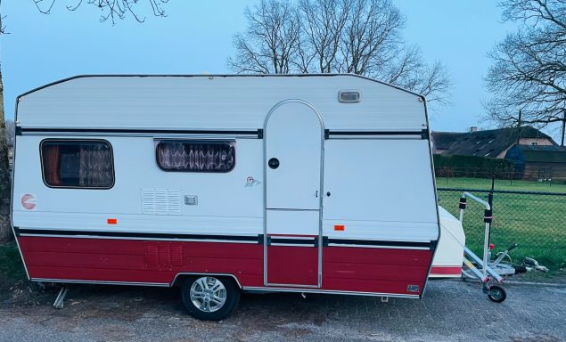 Witte en rode caravan met een deur aan de zijkant, geparkeerd op gras.